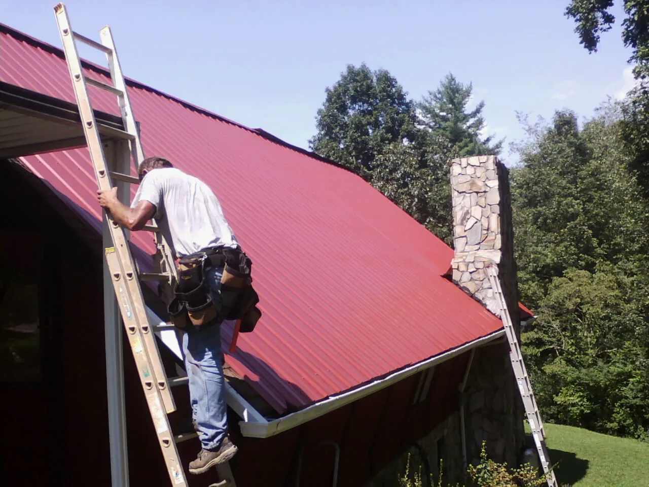 Roofer on a ladder beside a red metal roof and stone chimney — Soffit & Fascia Repair by Roofing Brothers in Bakersfield